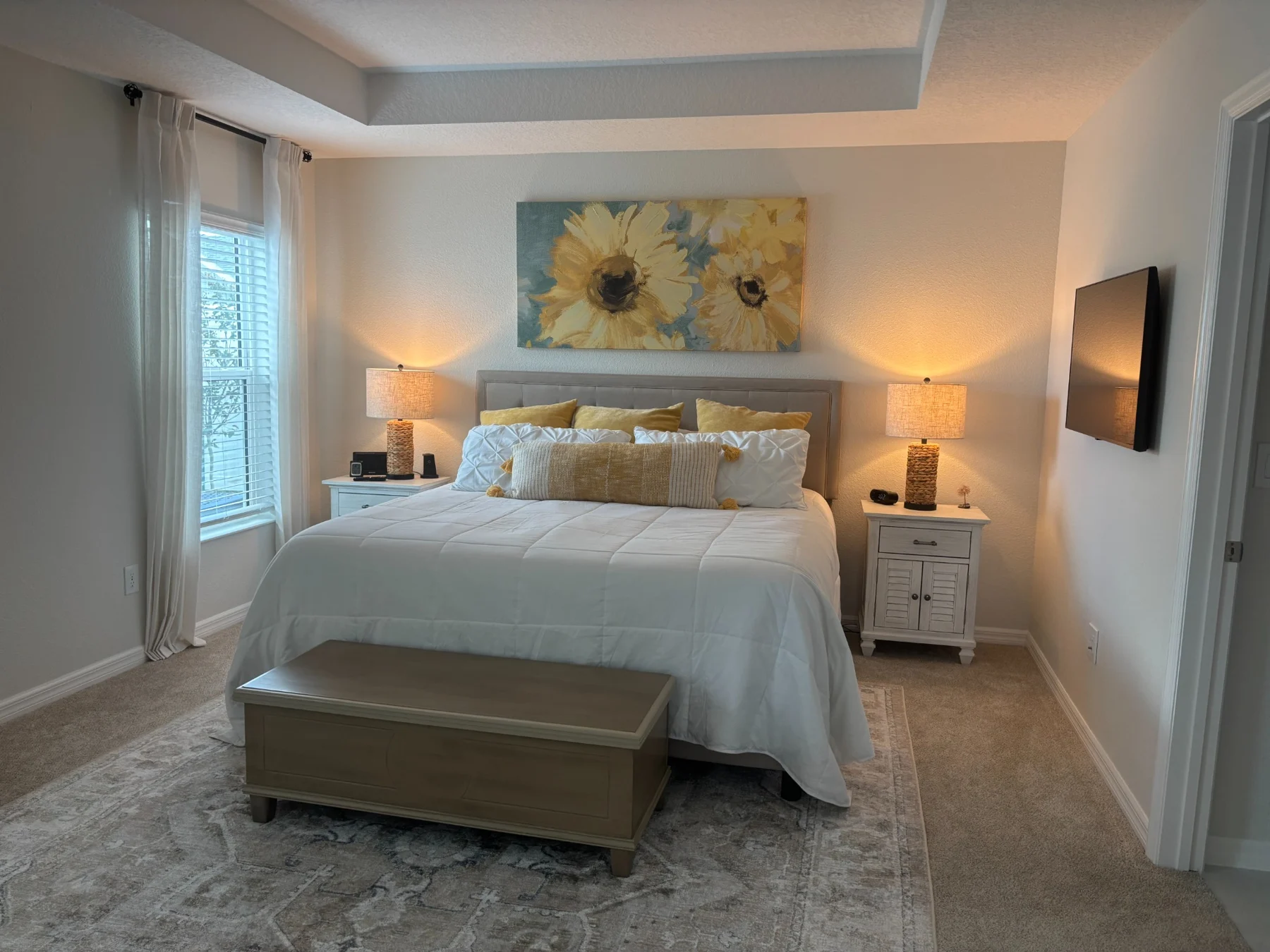 Bedroom with sunflower artwork, yellow accent pillows, white bedding, and a bench at the foot of the bed
